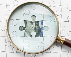 Magnifying glass hovering over paper money that’s hidden behind puzzle pieces 