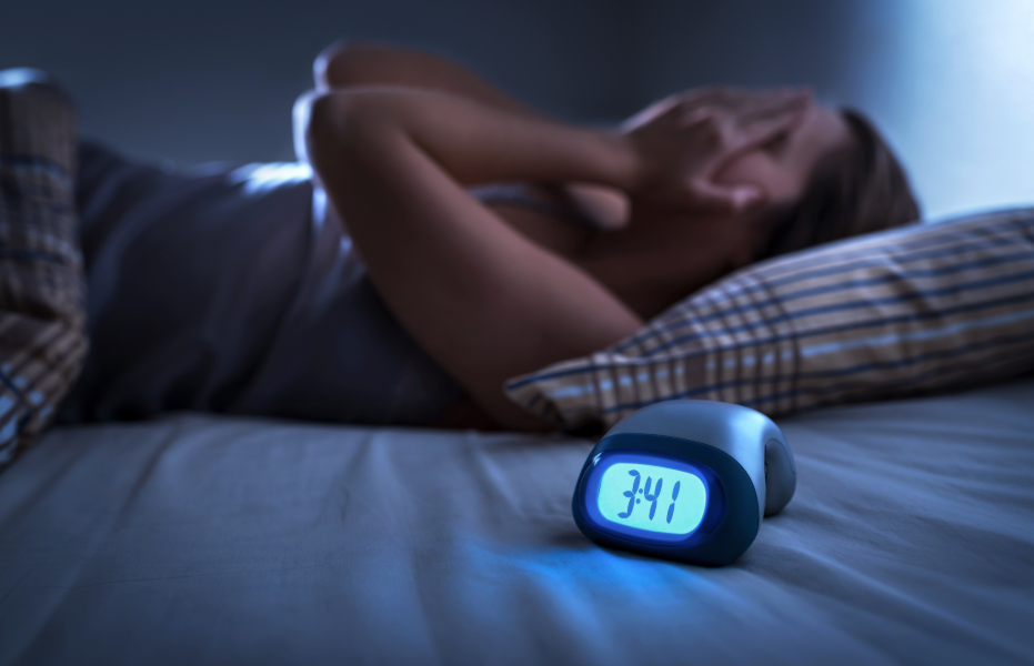 Woman covering face with hands, in bed, with alarm clock in foreground