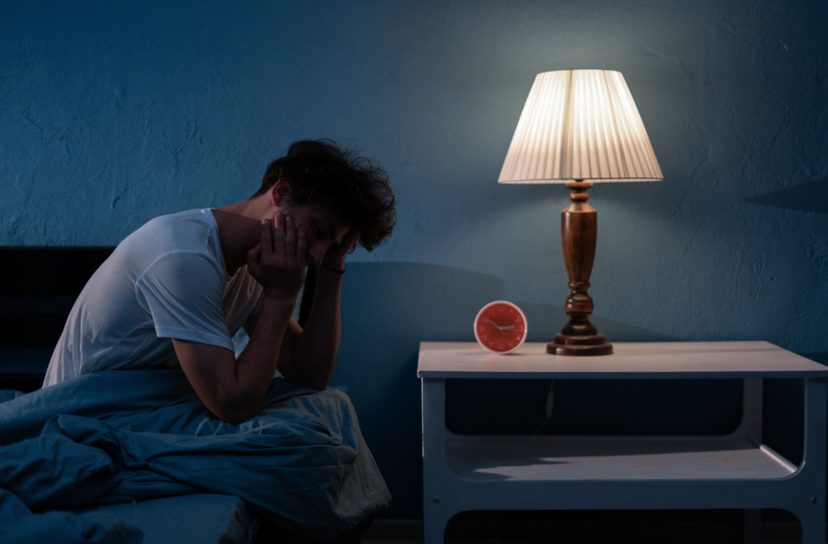 Man sitting on edge of bed with lamp on, holding head in hands, tired.